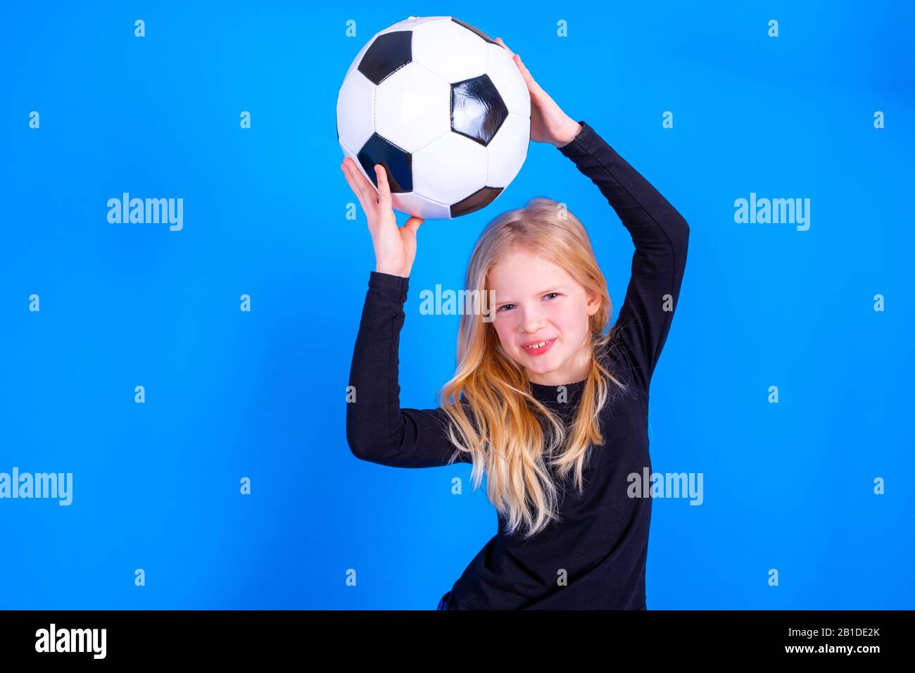 Tifosi di calcio. Cute bionda ragazza in camicia nera ha sollevato la palla sopra la sua testa su sfondo blu studio. Copyspace. Partita di calcio. Foto Stock