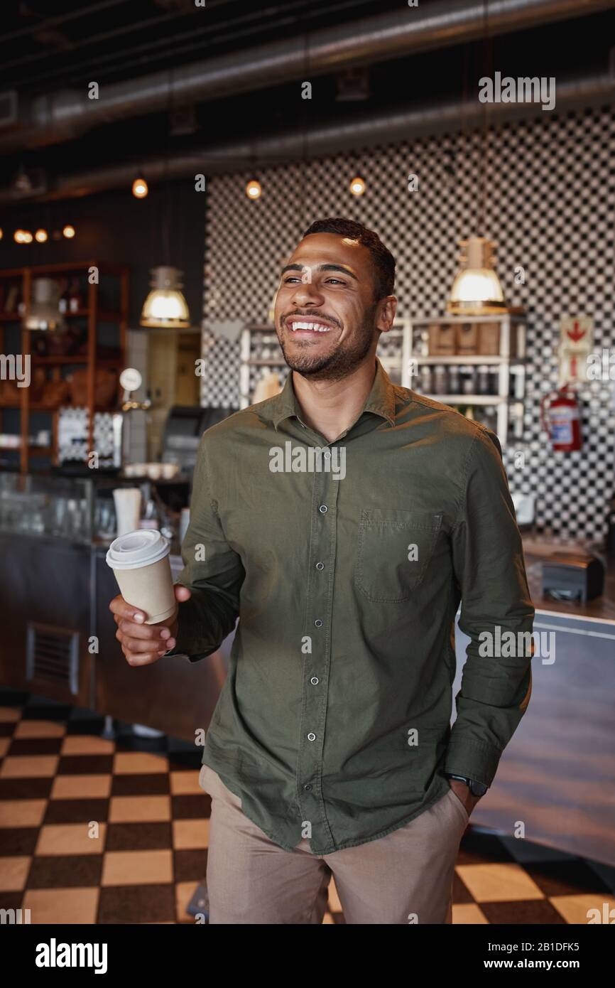 Bel giovane afro-americano che tiene la tazza del caffè del takeaway in piedi nel caffè Foto Stock