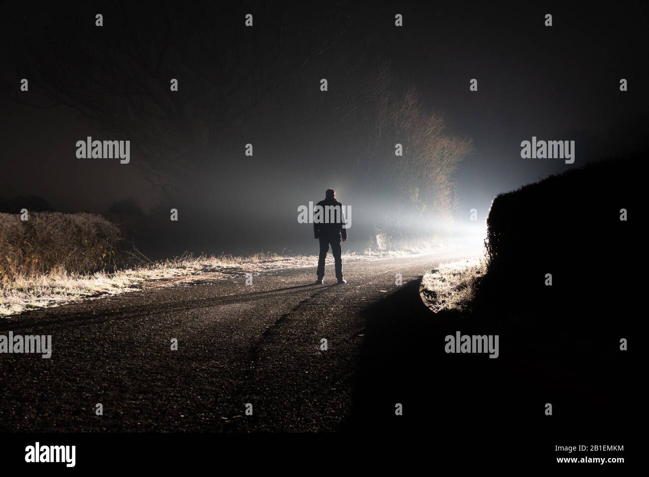 Una figura in piedi su una strada. Guardando una luce luminosa intorno a un angolo. Su una corsia di campagna spaventosa. In una notte di inverno misty. Foto Stock