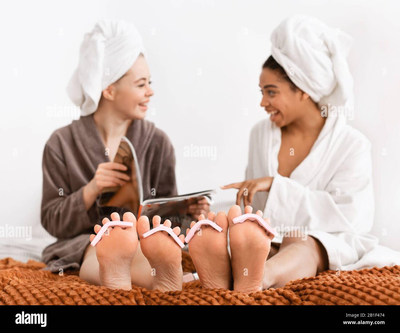Girlfriends in accappatoi facendo pedicure a casa, rivista di lettura Foto Stock