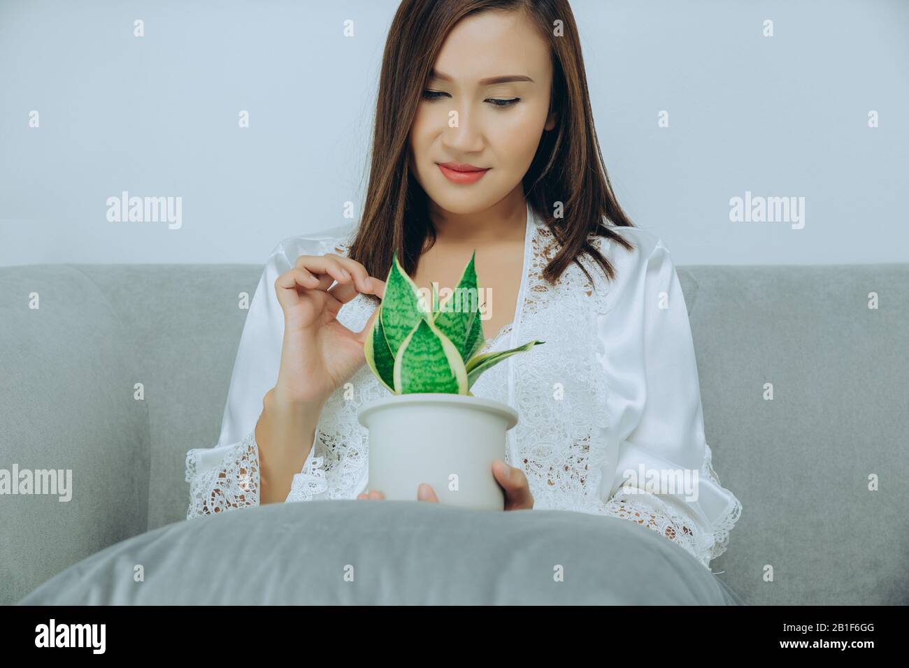 Le donne asiatiche indossano un abito da notte bianco e un lungo accappatoio in raso con laccio floreale mantengono le piante di purificazione dell'aria seduti sul divano grigio nella camera da letto al Nig Foto Stock