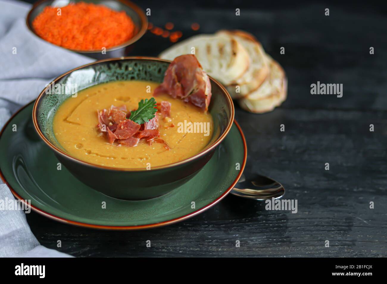 Zuppa di lenticchie su sfondo di legno. Servito con pancetta a fette ed erbe. Nelle vicinanze si trovano pezzi di ciabatta. Groats crudi in background. Spazio di copia Foto Stock