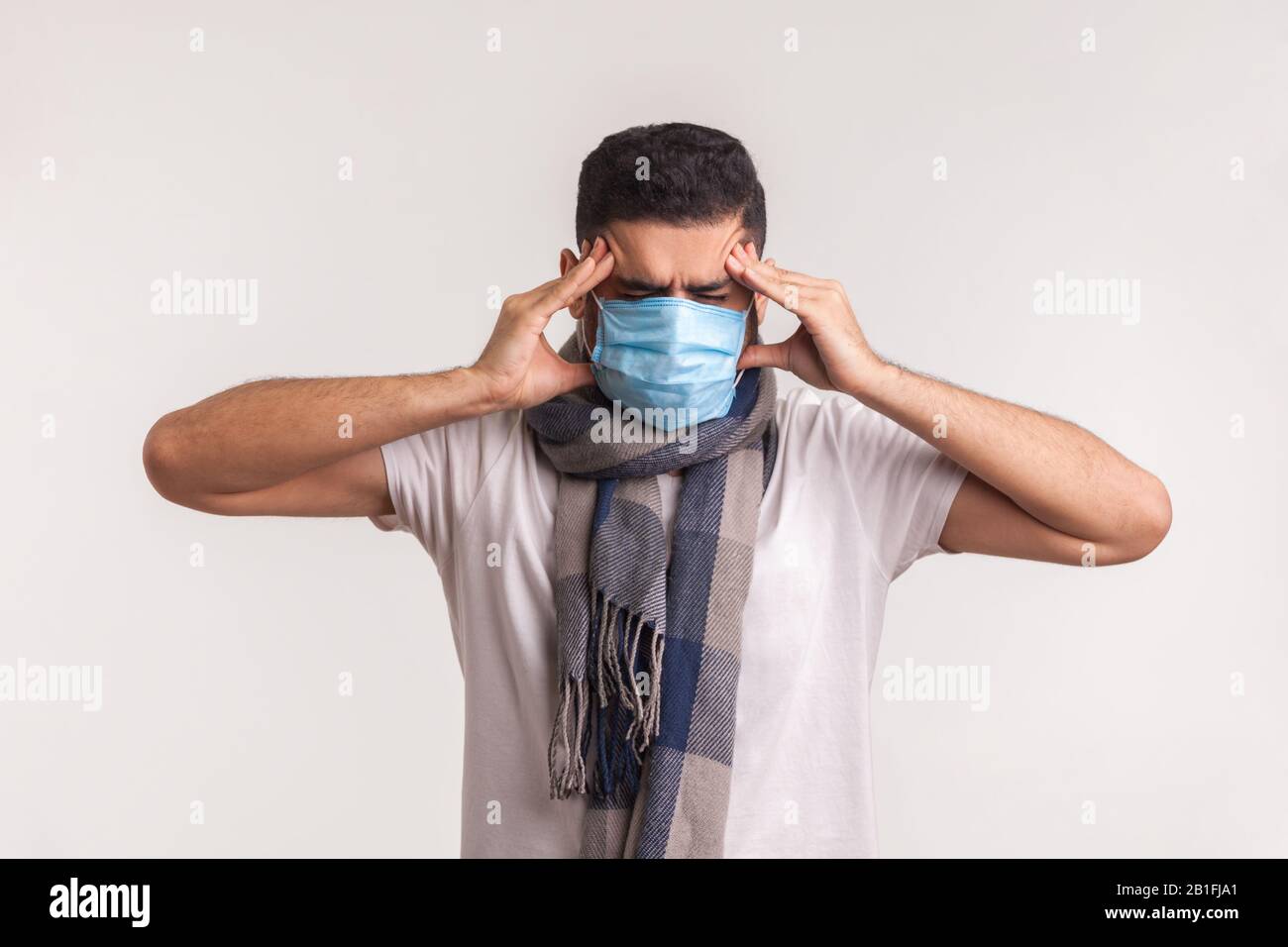 Uomo malsano in maschera chirurgica e sciarpa che soffre di mal di testa, rubbing templi dolorosi, avendo sintomi di influenza, contagiosa infezione da coronavirus 2019-nC Foto Stock