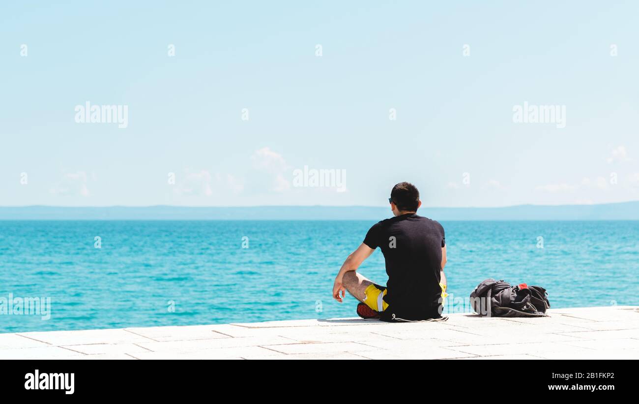 giovane uomo viaggiatore da solo con zaino vicino a lui meditando guardando l'oceano con copyspace Foto Stock
