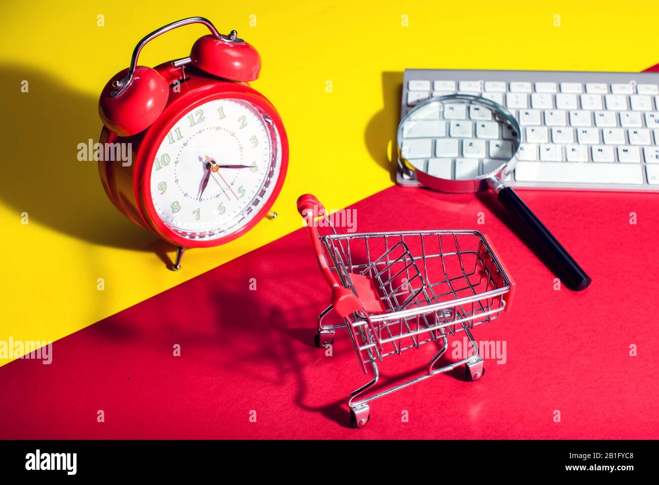 Sveglia rossa, piccolo carrello per lo shopping, tastiera e lente d'ingrandimento su sfondo a colori. Concetto di shopping Foto Stock