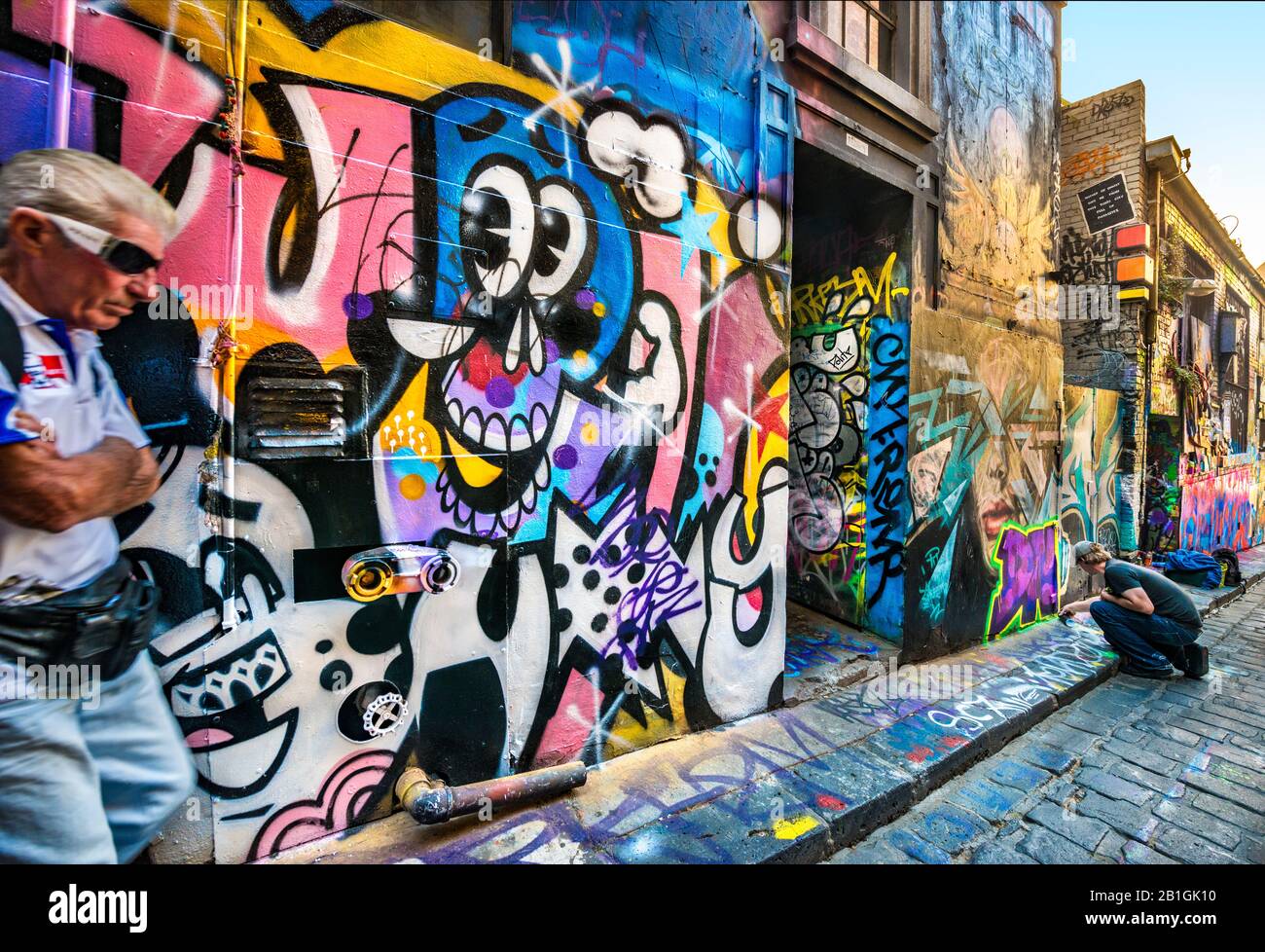 Uomo con occhiali da sole in piedi sul lato di strada acciottolata di vicolo di pietra, mentre giovane uomo spray dipinge graffiti, Hosier Street, Melbourne Lanes, Melbourne, Vic Foto Stock