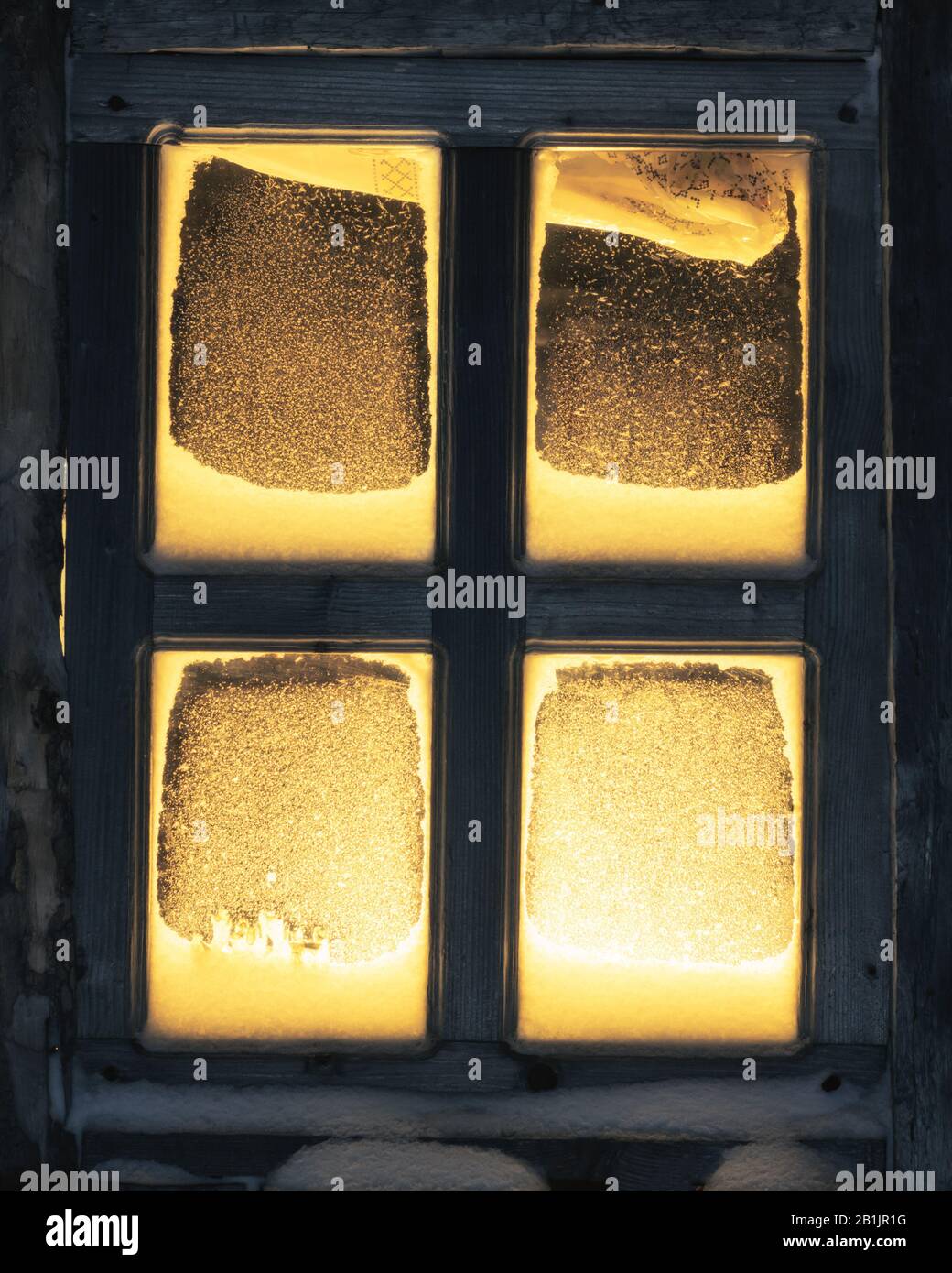 Finestra innevata su vecchia casa di legno con luce calda all'interno. Sera Foto Stock