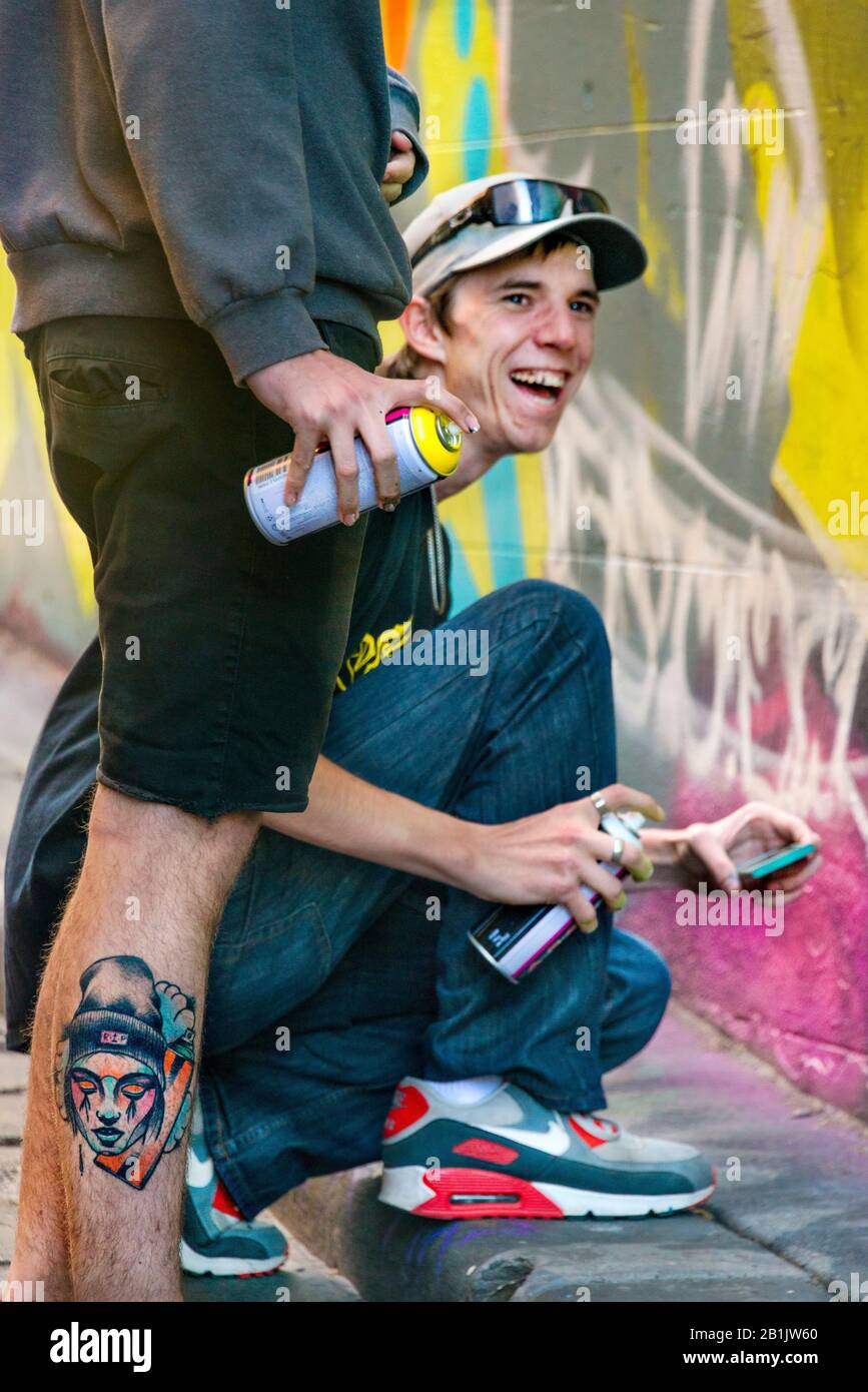 Giovane uomo felice, ridendo con il suo amico, come egli spray dipinge graffiti muro, Hosier Street, Melbourne Lanes, Melbourne, Victoria, Australia Foto Stock