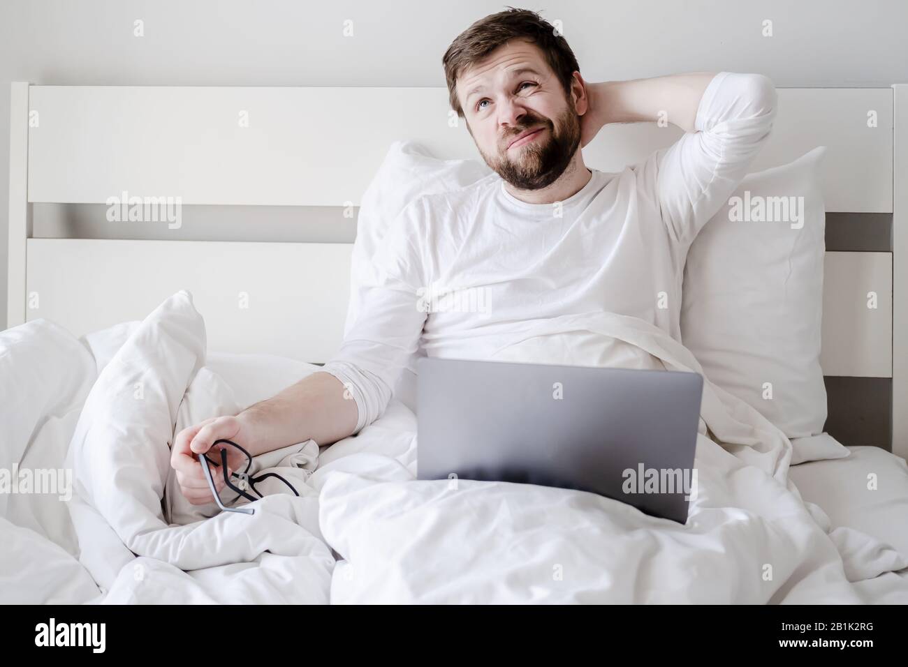 Blogger distratto dal lavorare con un computer portatile, ha tolto gli occhiali e guarda con attenzione, seduto a letto. Processo creativo. Foto Stock