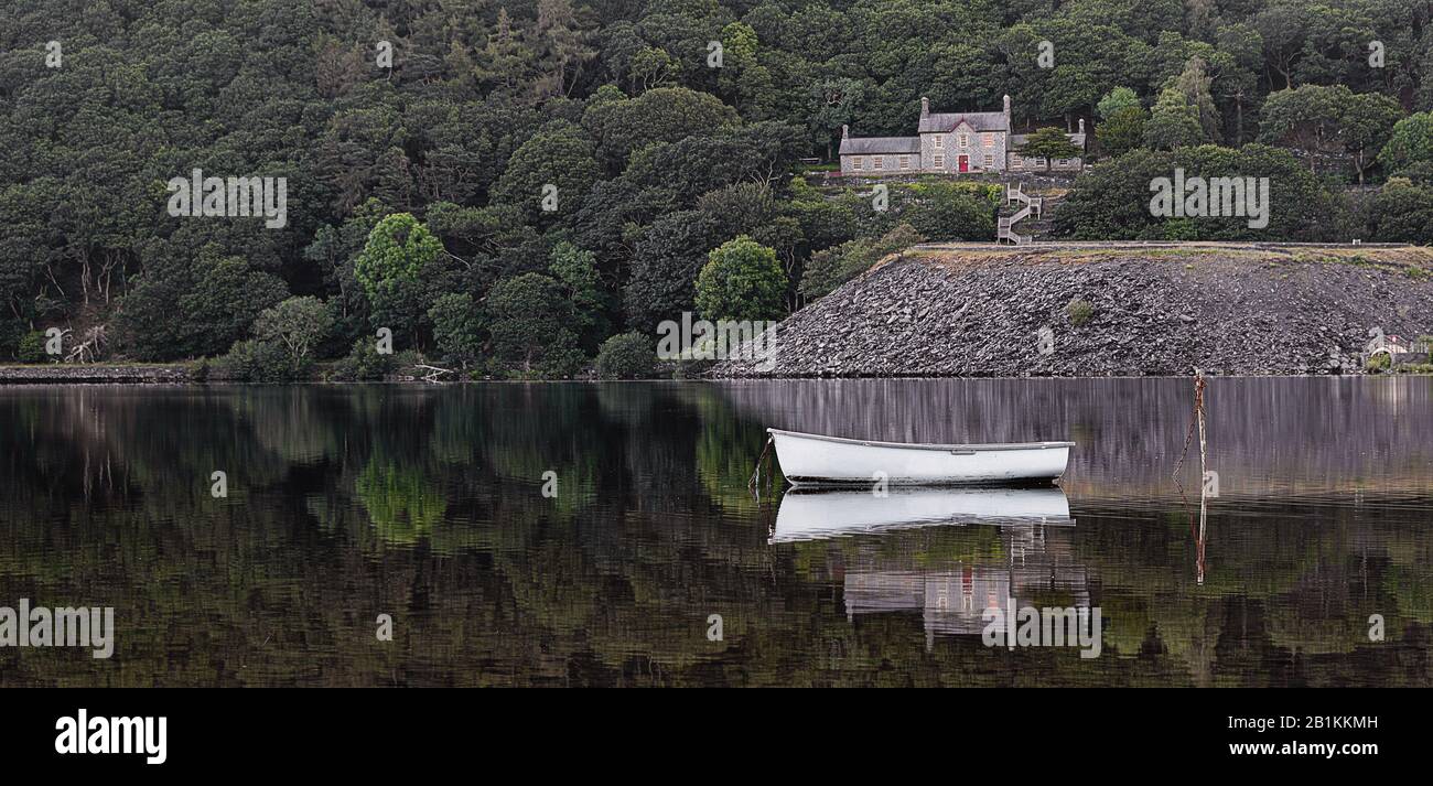 Una scena tranquilla su un lago. Una barca a remi è ormeggiata e c'è un edificio nel bosco dietro. Entrambi si riflettono nella calma acqua Foto Stock
