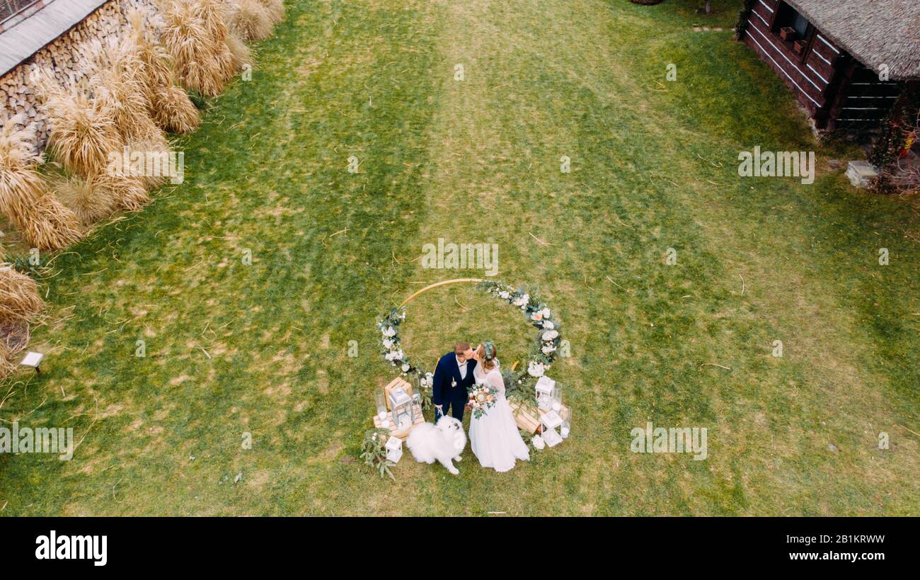 Vista dal drone dei newlyweds baciare sul prato accanto al cane sullo sfondo del matrimonio arco. Foto Stock