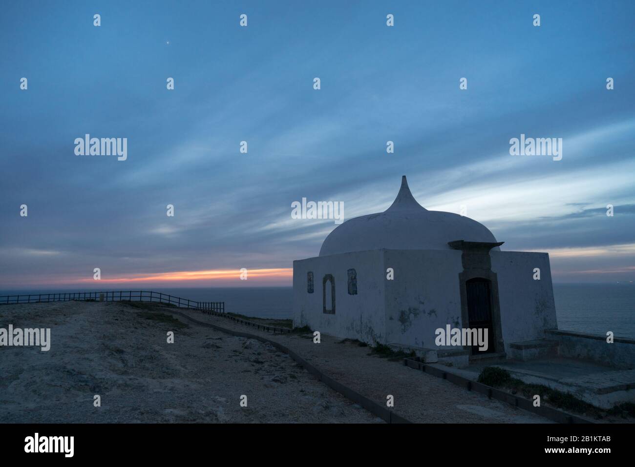 La cappella bianca di Ermida da Memória si trova ai margini di una scogliera vicino all'Oceano Atlantico al tramonto con un cielo nuvoloso e pantone classico blu Foto Stock