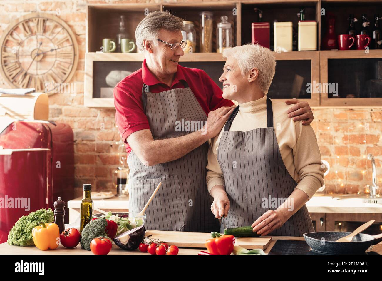 Amorevole coppia senior indossando grembiuli e abbracciando in cucina Foto Stock