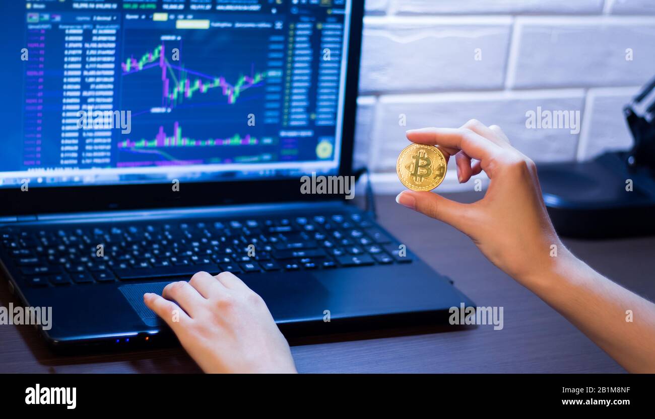 Criptovaluta. Una ragazza tiene una moneta di Bitcoin seduta davanti ad un  laptop che analizza il