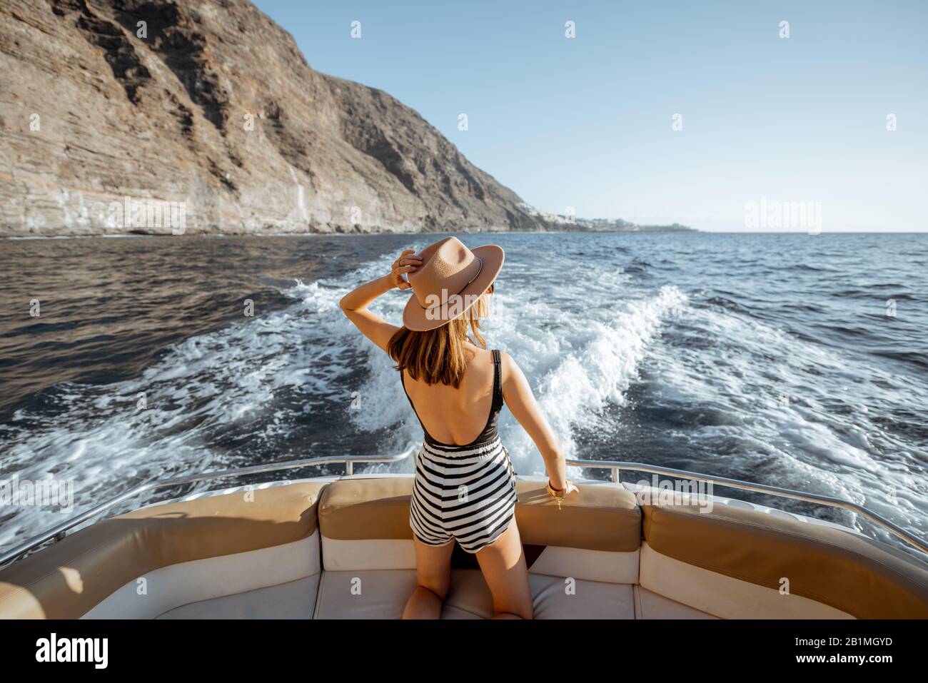 Rilassante attraente donna in cappello sole godendo viaggio oceano, navigando sul yacht di lusso vicino alla splendida costa rocciosa su un tramonto. Vista dal retro Foto Stock