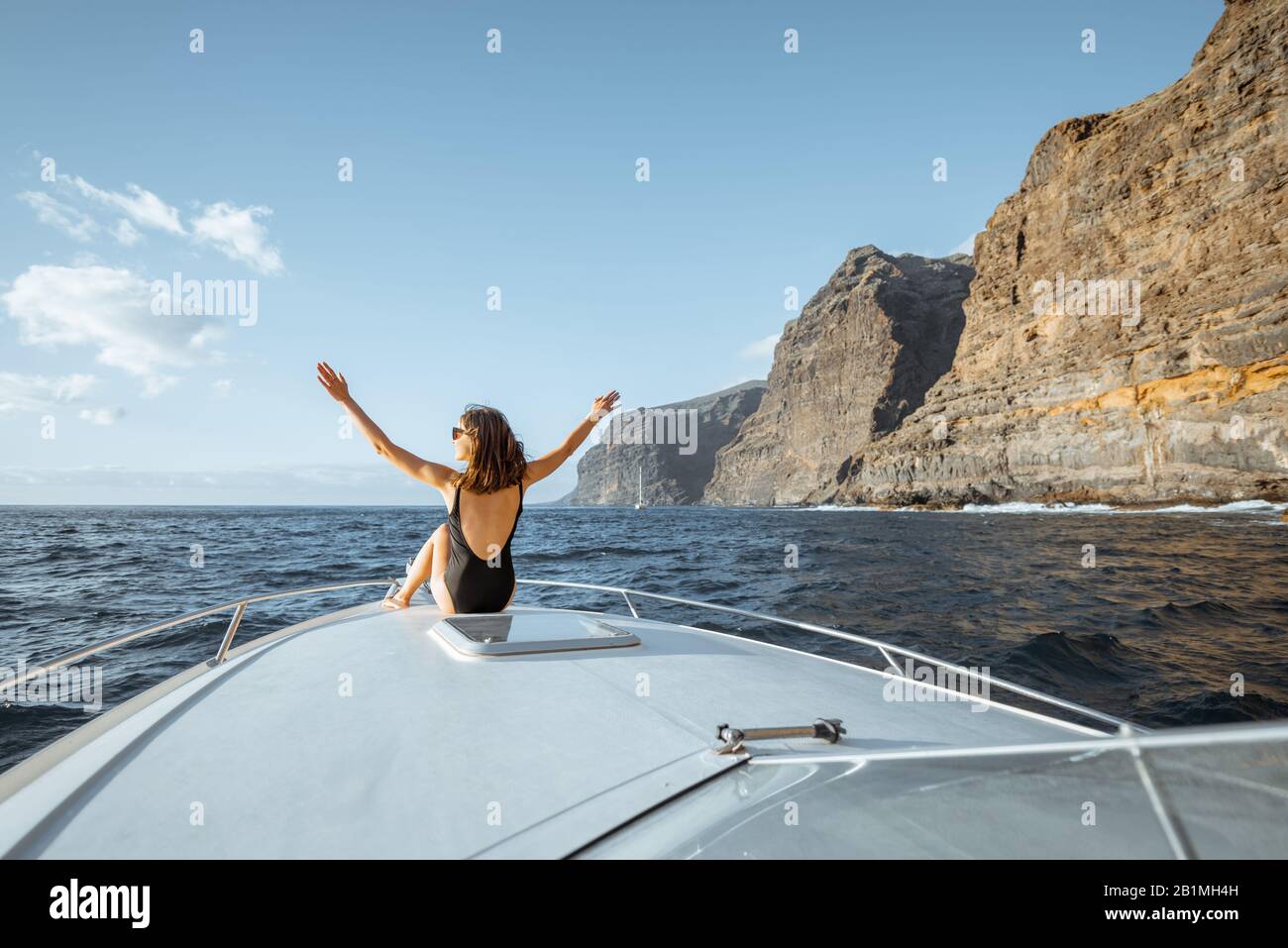 Donna che gode di un viaggio in oceano seduto sul naso dello yacht mentre naviga vicino alla costa rocciosa mozzafiato su un tramonto Foto Stock