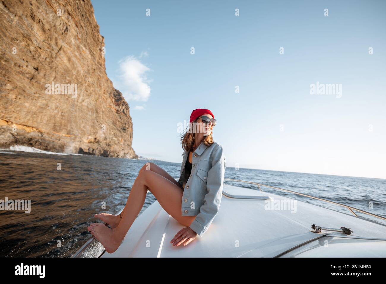 Donna vestita casualmente godendo viaggio oceanico, navigando su uno yacht vicino alla costa rocciosa mozzafiato su un tramonto. Concetto di uno stile di vita e di viaggio spensierati Foto Stock