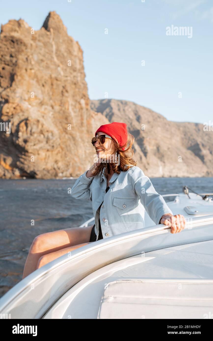 Donna vestita casualmente godendo viaggio oceanico, navigando su uno yacht vicino alla costa rocciosa mozzafiato su un tramonto. Concetto di uno stile di vita e di viaggio spensierati Foto Stock