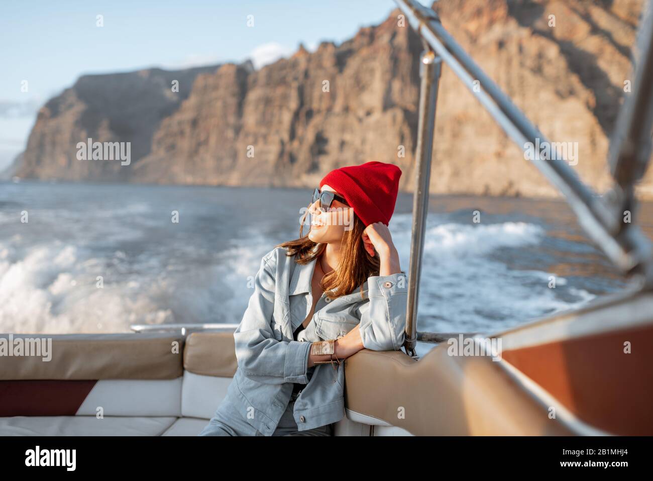 Ritratto di una giovane viaggiatore vestito casualmente con cappello rosso e jeans che naviga su uno yacht vicino alla costa rocciosa. Concetto di uno stile di vita e di viaggio spensierati Foto Stock