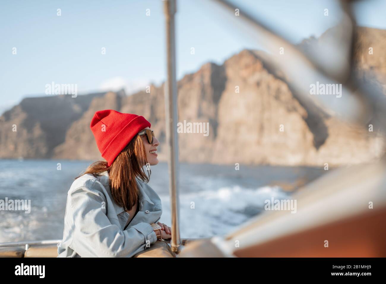 Ritratto di una giovane viaggiatore vestito casualmente con cappello rosso e jeans che naviga su uno yacht vicino alla costa rocciosa. Concetto di uno stile di vita e di viaggio spensierati Foto Stock