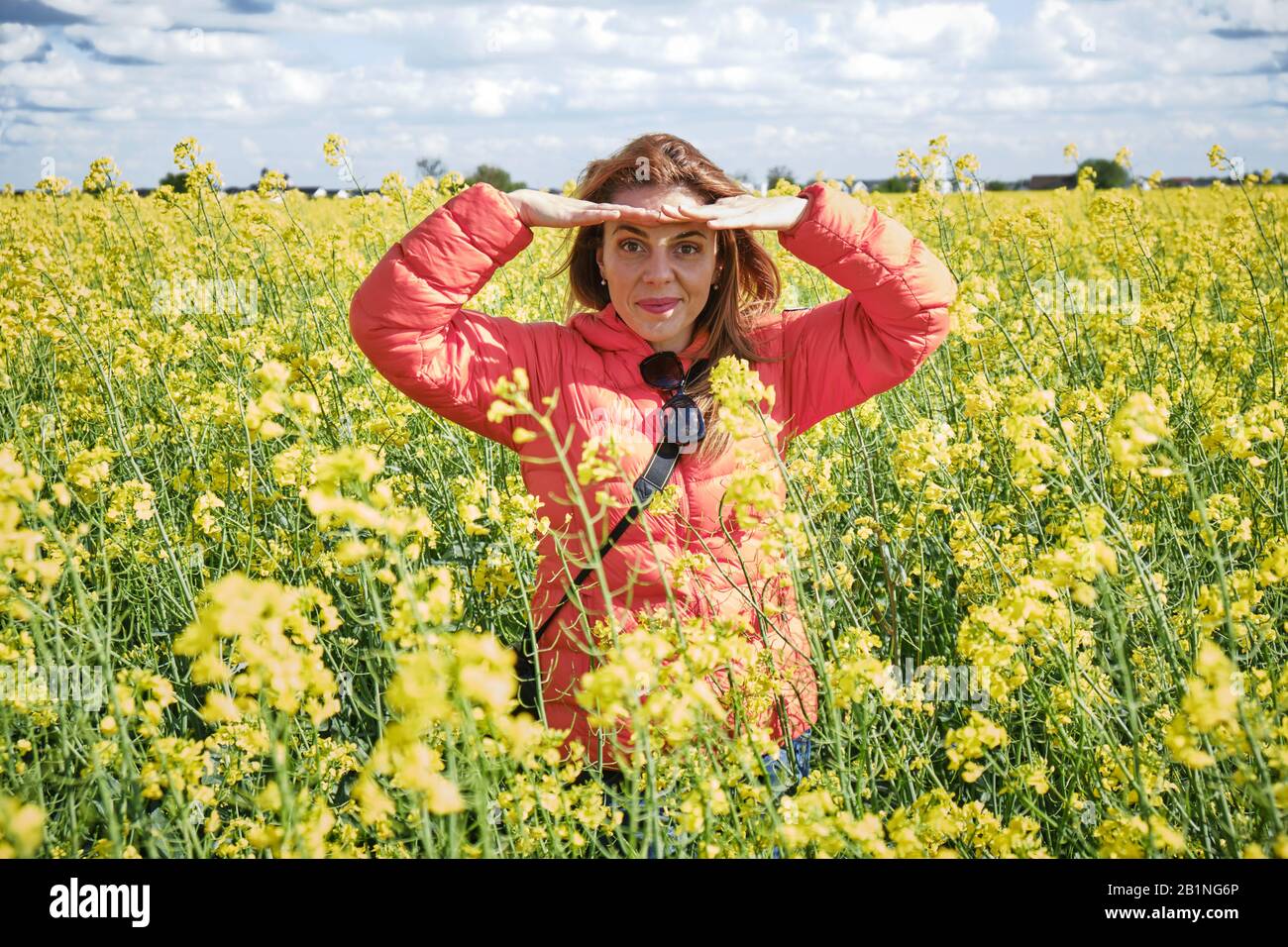 Bella donna in un campo di colza che ombreggiano gli occhi dal sole luminoso, usando entrambe le mani per bloccare la luce del sole, mentre guardando dritto verso il Foto Stock