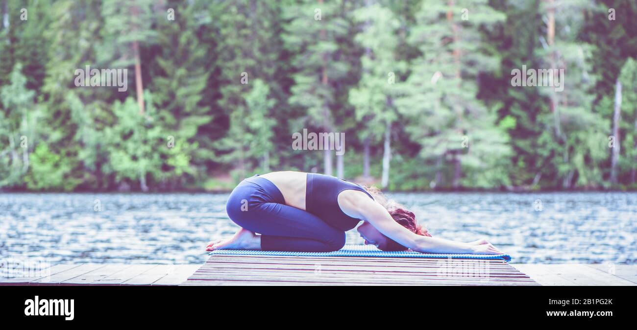Giovane ragazza attraente pratica yoga, seduta in esercizio di bambino, Balasana posa sul lago. Il concetto di pacificazione, stile di vita sano. Panorami Foto Stock