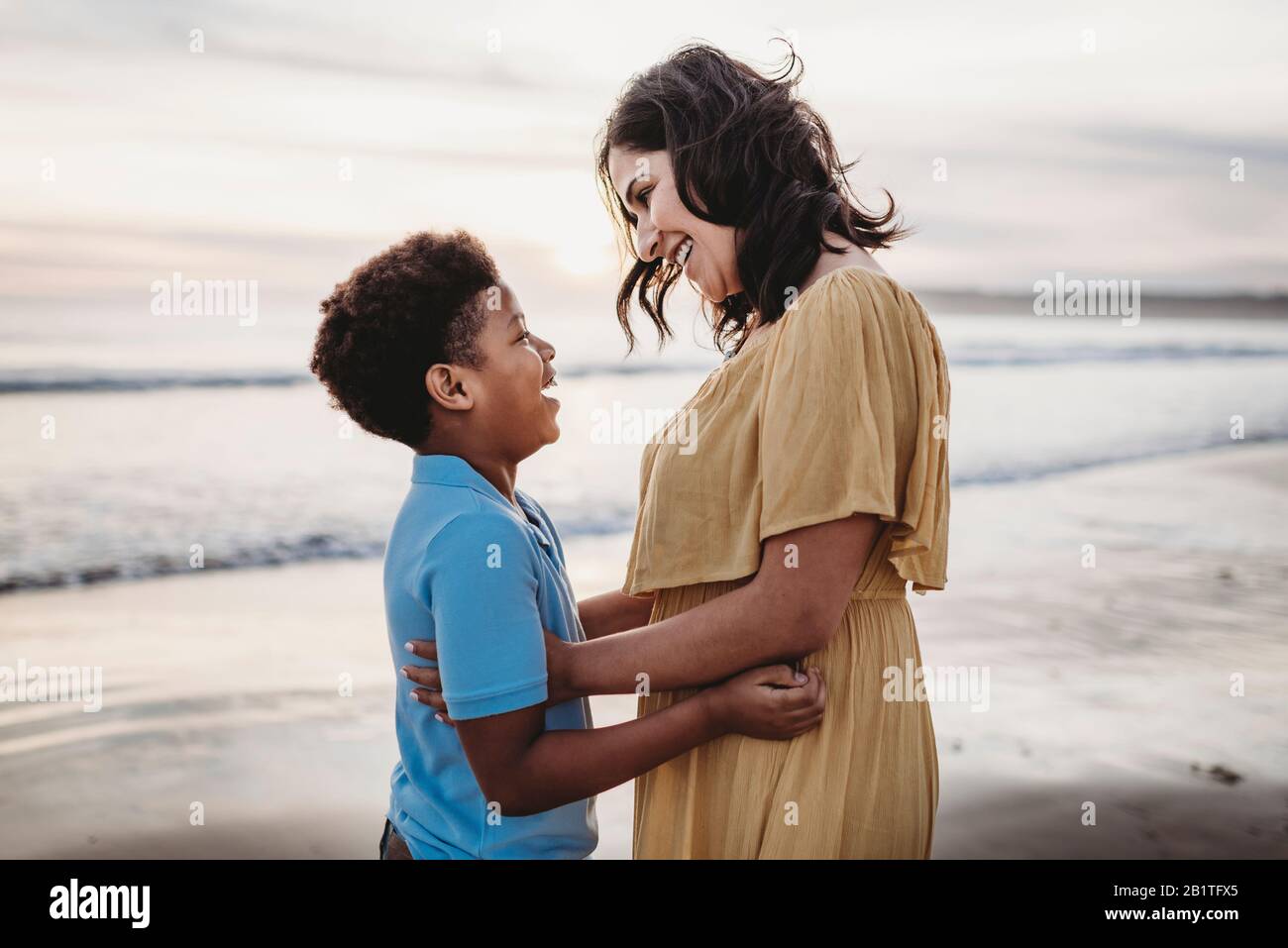 Vista laterale della bella madre che abbraccia il figlio al mare durante il tramonto Foto Stock