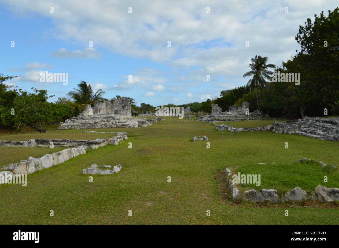 Un'ampia vista panoramica delle antiche rovine maya in pietra a Tulum, Messico, con pareti intemprate, colonne, erba verde, alberi tropicali e una b parzialmente nuvolosa Foto Stock