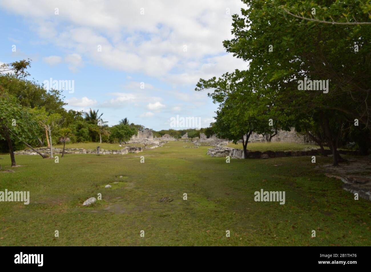 Un'ampia vista panoramica delle antiche rovine maya in pietra a Tulum, Messico, con pareti intemprate, colonne, erba verde, alberi tropicali e una b parzialmente nuvolosa Foto Stock