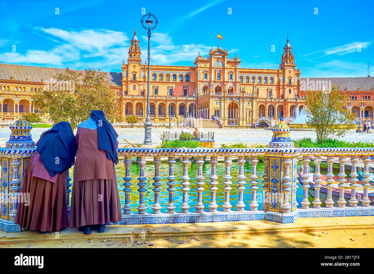 Due monache poggiano su mancorrenti in ceramica del canale sotto l'ombra degli alberi e osservano l'attività della gente su Plaza de Espana, Siviglia, Spagna Foto Stock
