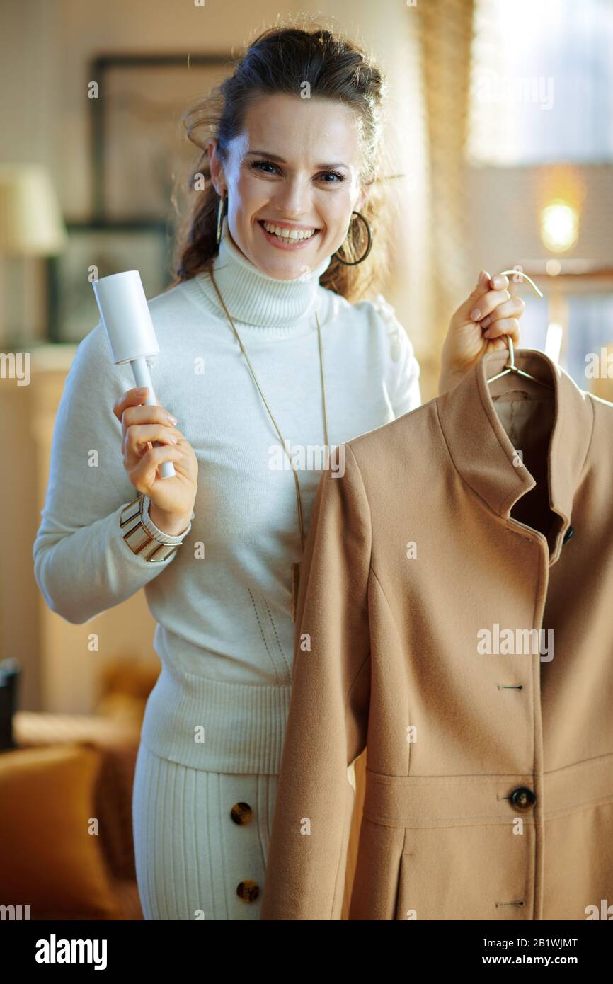 Preparazione per la stagione fredda. Sorridendo moderna casalinga di 40 anni in maglione bianco e mantello cappotto di pulizia su appendiabiti con rullo di pelucchi nel moderno hou Foto Stock