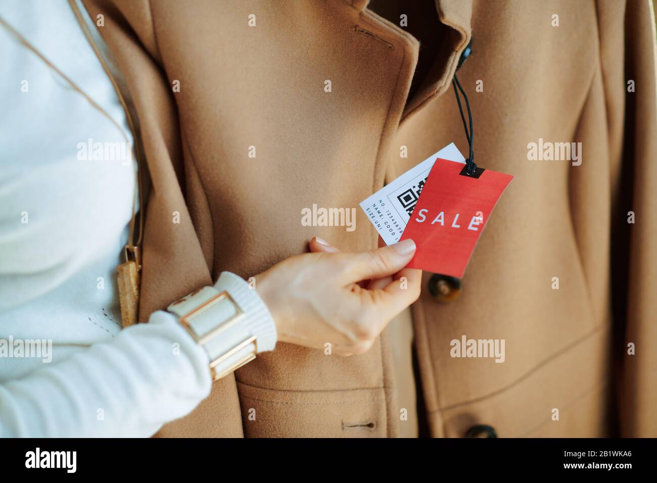 Closeup su donna in maglione bianco e gonna in negozio di moda moderno esamina il prezzo di vendita tag su cappotto beige. Foto Stock