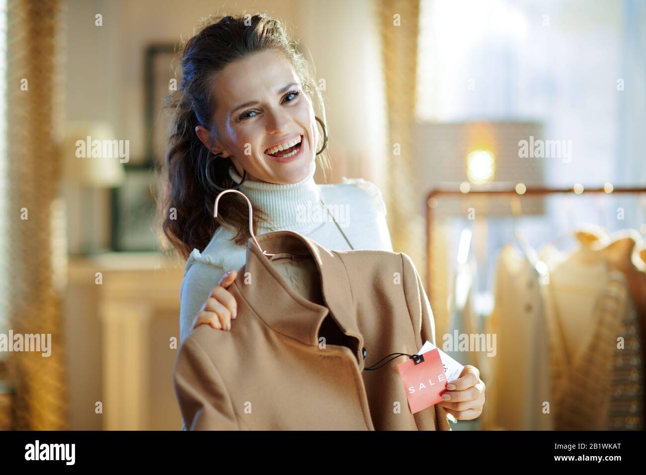 felice donna elegante in maglione bianco e gonna che mostra cappotto beige con prezzo di vendita tag in negozio di moda moderno. Foto Stock