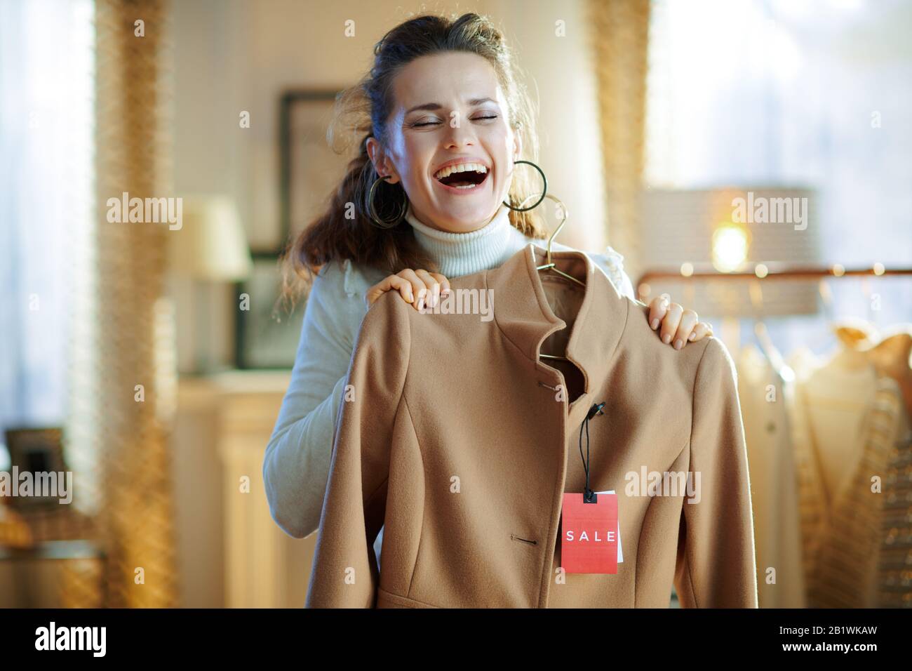 donna sorridente di 40 anni in maglione bianco e gonna che tiene cappotto beige con prezzo di vendita tag in moderno showroom moda. Foto Stock
