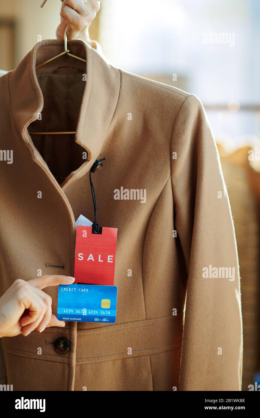 Primo piano su donna che tiene cappotto beige con prezzo di vendita tag su appendiabiti e carta di credito blu in moderno showroom moda. Foto Stock