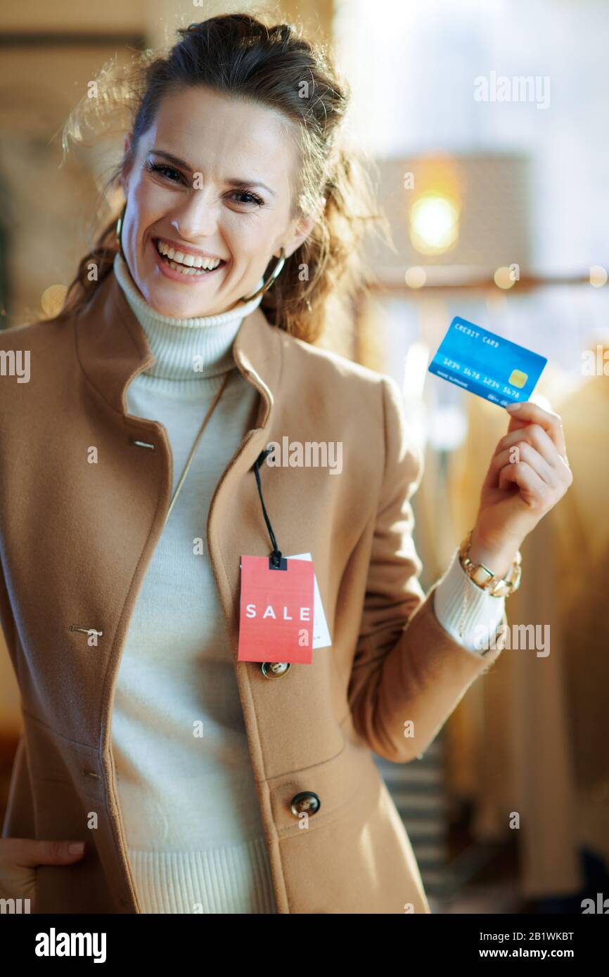 felice donna elegante in maglione bianco e gonna cercando cappotto beige con prezzo di vendita rosso tag e tenendo carta di credito blu in moderno showroom moda. Foto Stock