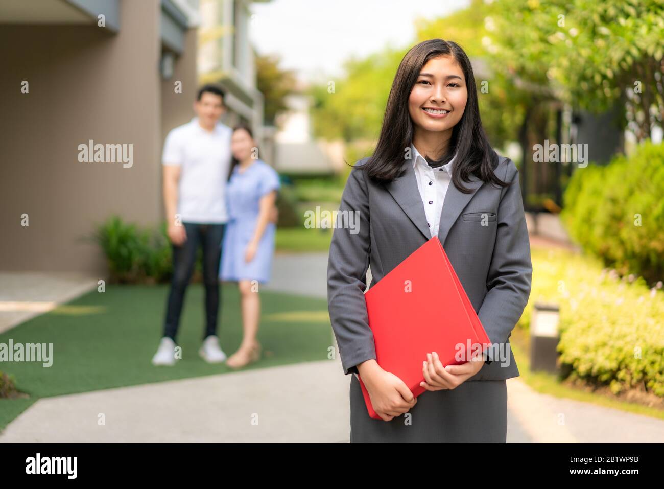 Asian fiducioso donna agente immobiliare o agente immobiliare in tuta tenendo file rosso e sorriso con giovane coppia venditori casa dietro di fronte a casa. Portrai Foto Stock