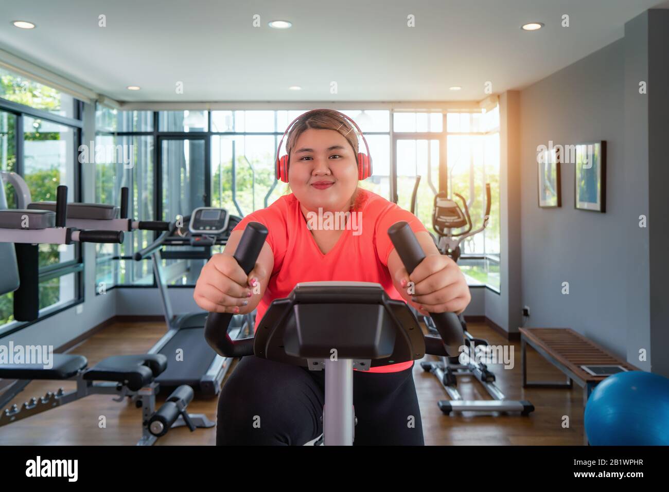 Felice Asian Overweight donna con l'allenamento degli auricolari su cyclette in palestra moderna, felice e sorridente durante l'allenamento. Donna grassa prendersi cura di salute e. Foto Stock