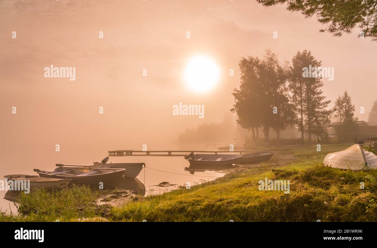 Bel sole dorato si illumina attraverso la nebbia al lago durante l'alba precoce, barche e lungo ponte di legno, alberi, riflesso cielo in acqua calma, spazio copia Foto Stock