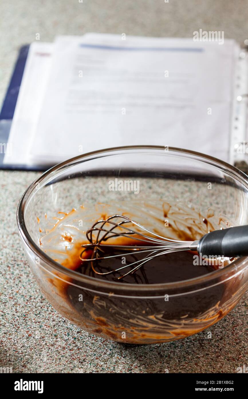 cioccolato fuso in una ciotola di vetro con una frusta e un libro aperto di ricetta fuori fuoco Foto Stock