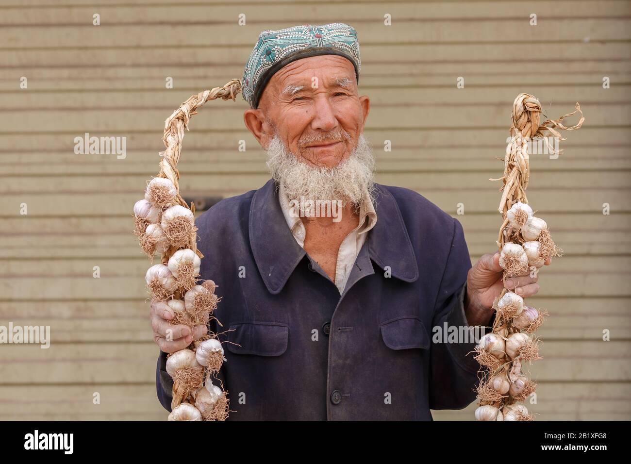 Uomo anziano uyghur con barba grigia che tiene corde di aglio in un bazar a Kashgar. L'uomo indossa un tradizionale cappello doppa. Foto Stock