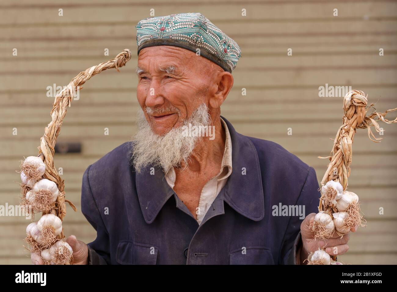 Sorridente, anziano uyghur uomo con una testa doppa che tiene due corde di aglio ad un mercato a Kashgar. Foto Stock