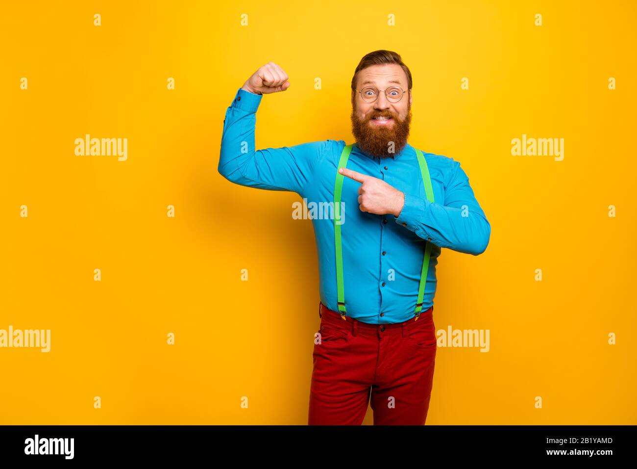 Guarda. Foto di attraente divertente ragazzo toothy sollevare pugno braccio che indica il dito che presenta forma perfetta bicipite indossare camicia blu verde sospenders pantaloni rossi Foto Stock