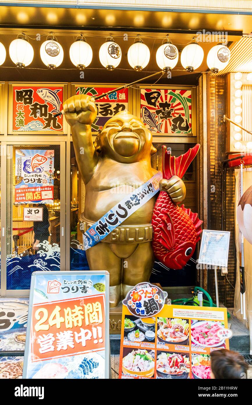 Statua di Billiken, icona del quartiere di Shinsekai, Osaka, che tiene i pesci in piedi lungo il sentiero e braccio sollevato reggendo la canna da pesca, al di fuori del ristorante di pesce. Foto Stock
