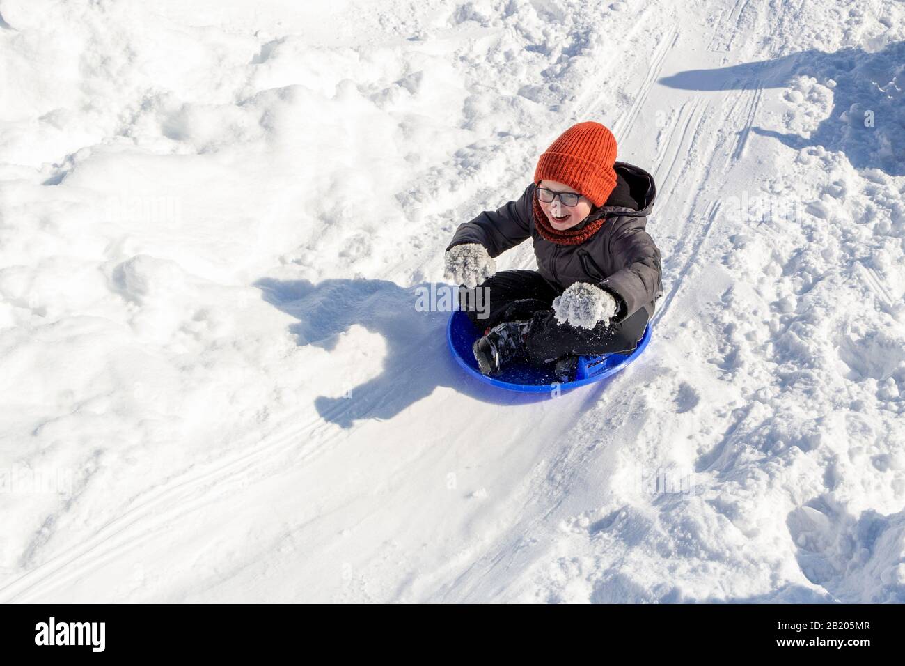 Divertimento per i bambini in inverno. Slittino da uno scivolo sulla neve.  Un ragazzo sorridente in occhiali si muove in una slitta, come un pilota  come se guidasse un cavallo redini. Divertimento
