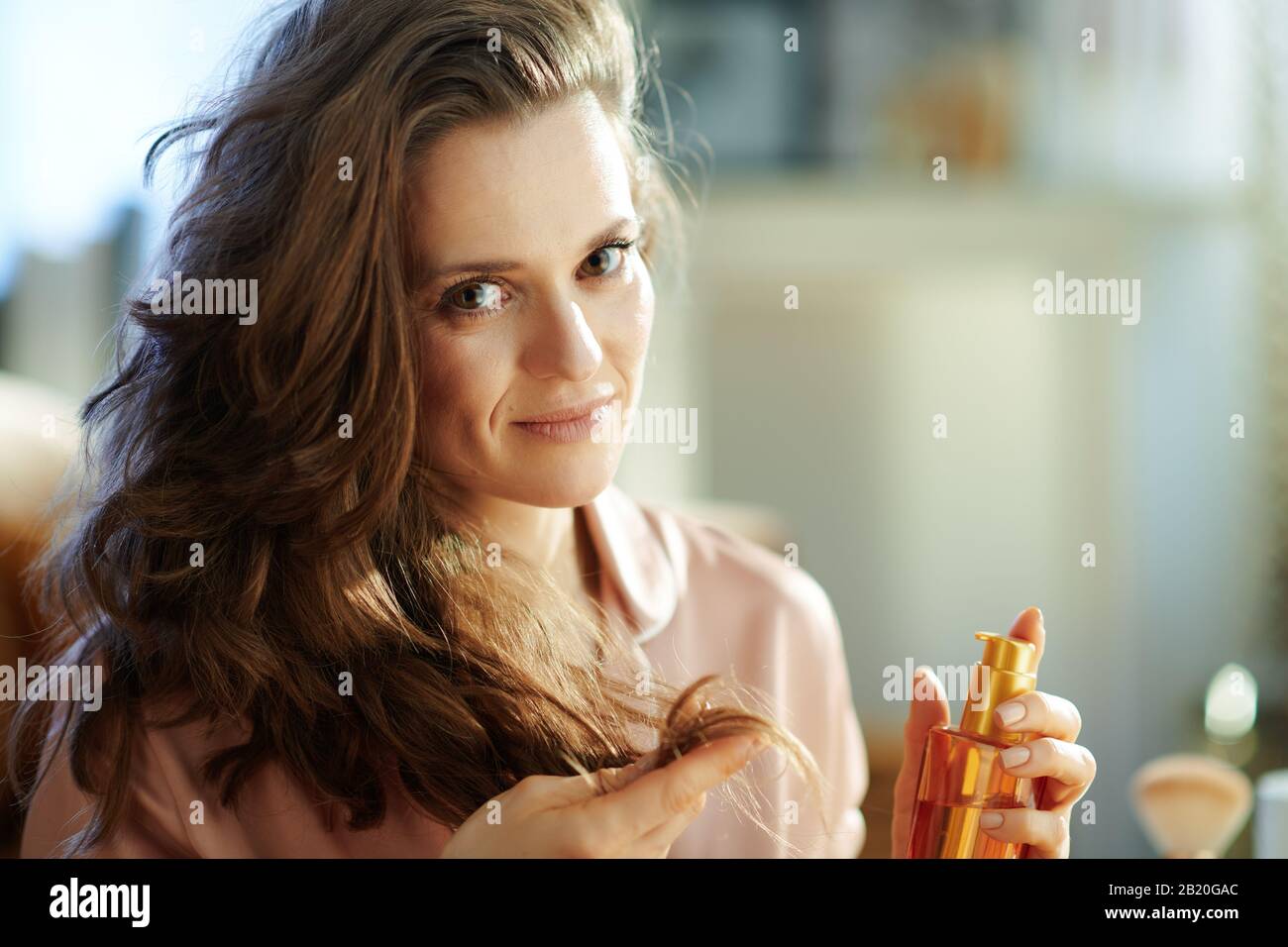 Ritratto di moderna donna di mezza età in pigiami con lungo brunette capelli ondulati tenere bottiglia di olio per capelli e controllare le estremità dei capelli nel moderno soggiorno i Foto Stock