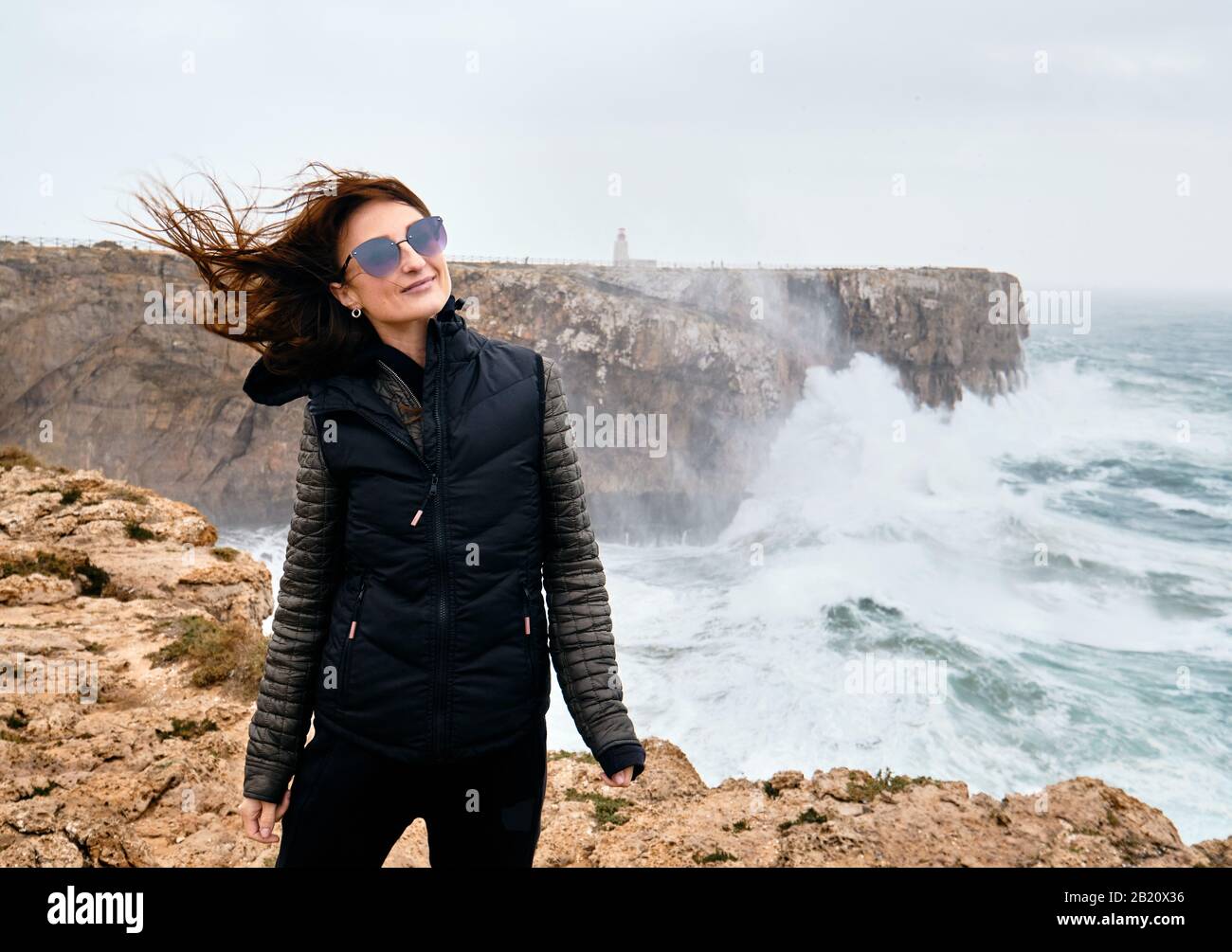 Millennial caucasica donna i suoi capelli fluttering nel vento che si posa in piedi sul bordo della scogliera, tempestoso Oceano Atlantico sullo sfondo. Fortaleza Sagres Foto Stock