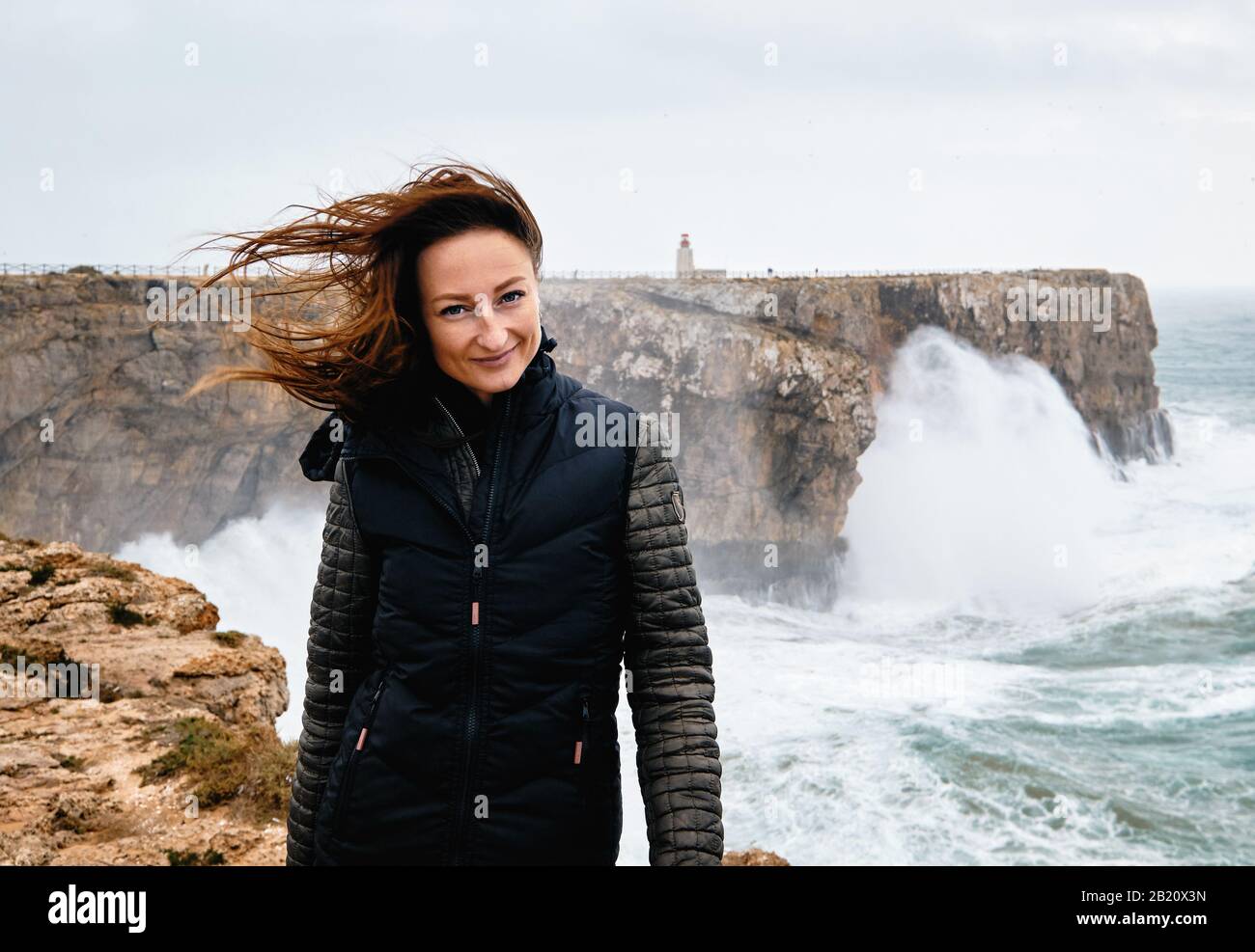 Millennial caucasica donna i suoi capelli fluttering nel vento che posa guardando la macchina fotografica in piedi sul bordo della scogliera, stormy Oceano Atlantico sullo sfondo Foto Stock