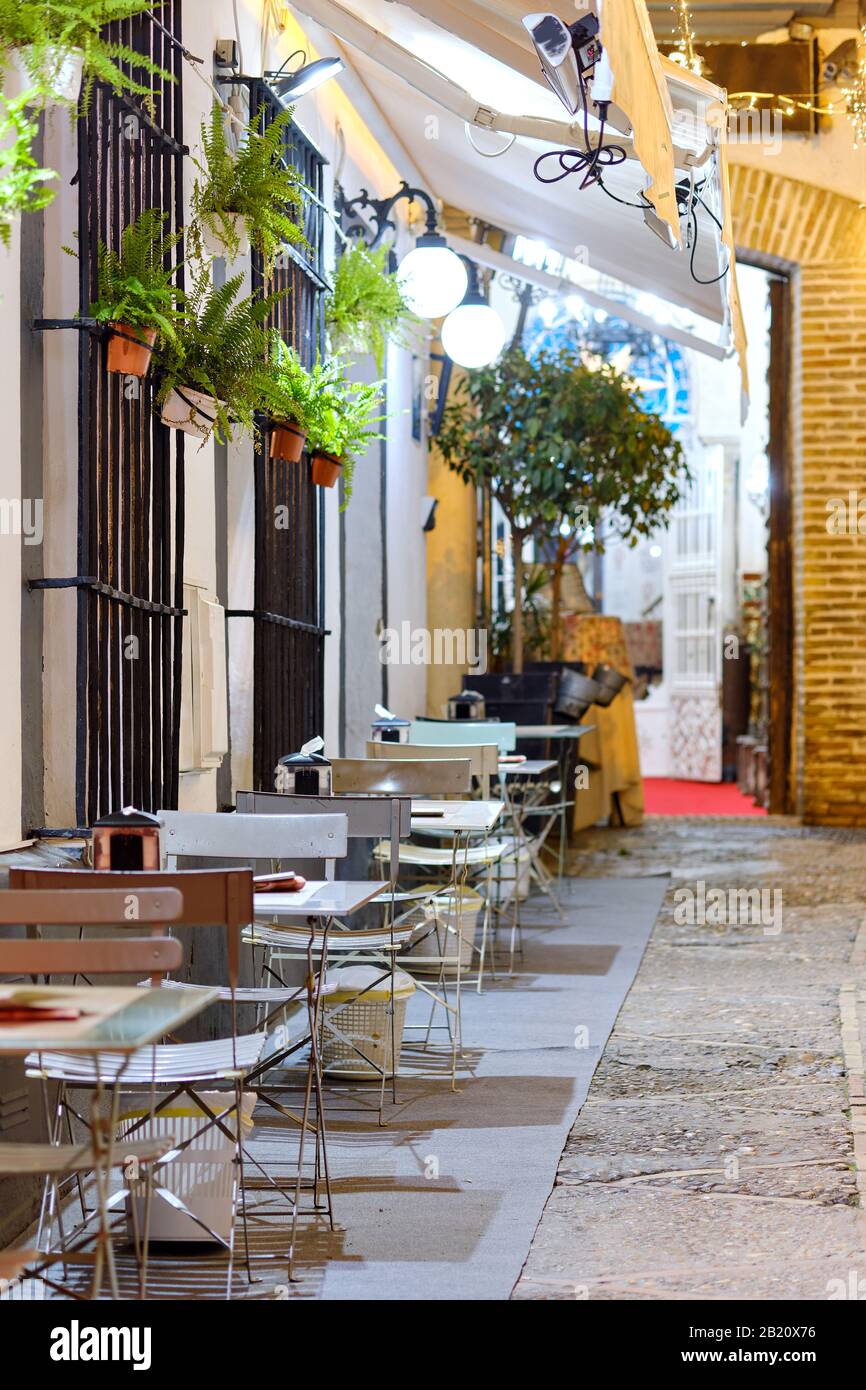 Affascinante marciapiede terrazzato open space cafe, vuoti tavoli metallici e sedie nella strada illuminata da lanterne vista notturna della città spagnola di Siviglia. Spagna Foto Stock