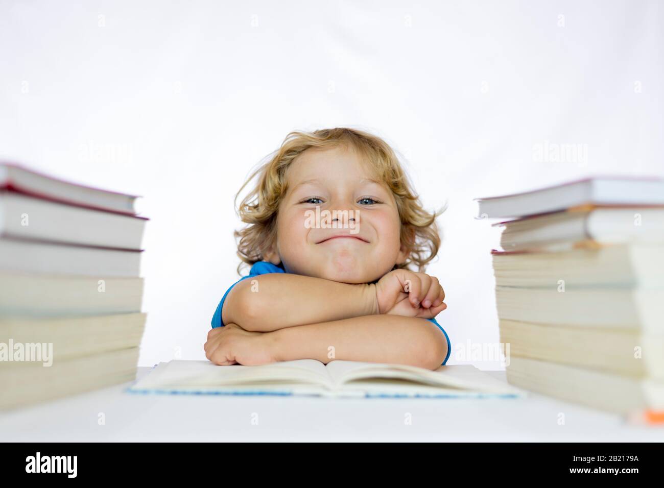 Piccolo bambino felice tra i 4 e i 5 anni seduto ad una scrivania che studia con i libri di testo sul suo tavolo e con uno sfondo bianco Foto Stock